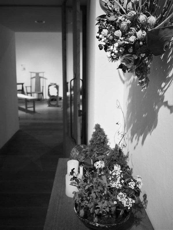 前景にドライフラワーのアレンジメント、背景にぼやけた廊下と木製家具の展示が続くモノクロ写真